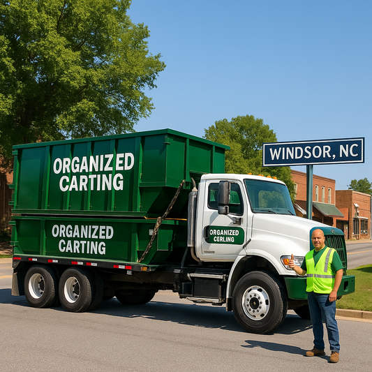A green and white Organized Carting flatbed truck hauling two large roll-off dumpsters on a sunny day in downtown Windsor, North Carolina, with the driver in a neon safety vest giving a thumbs-up next to the “Windsor, NC” sign.